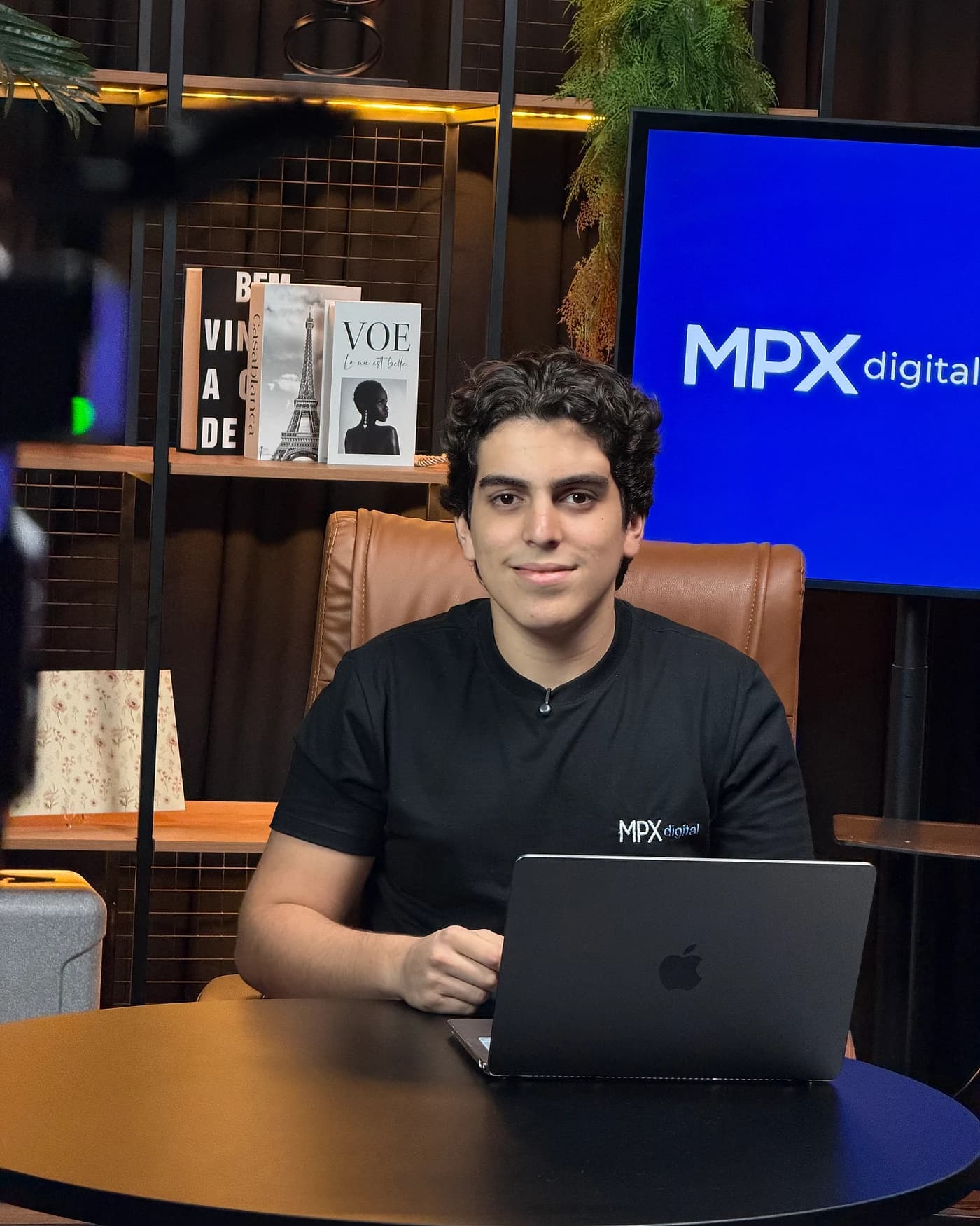 Mateus Paculdino, da MPX Digital, trabalhando no notebook no estúdio da empresa, com a marca MPX Digital exibida no telão ao fundo