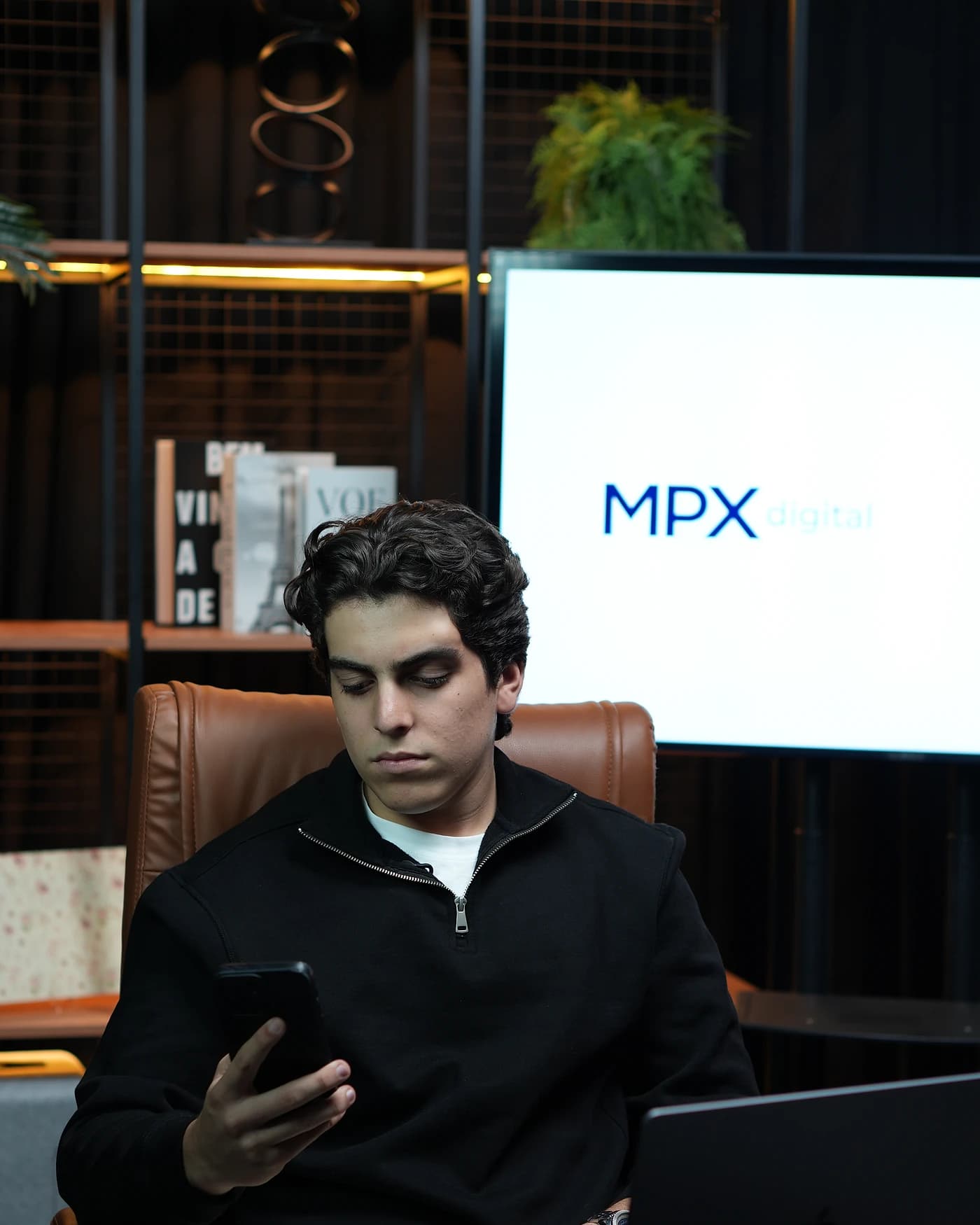 Mateus Paculdino em trabalho no estúdio da MPX Digital, concentrado no notebook, com a marca no telão ao fundo